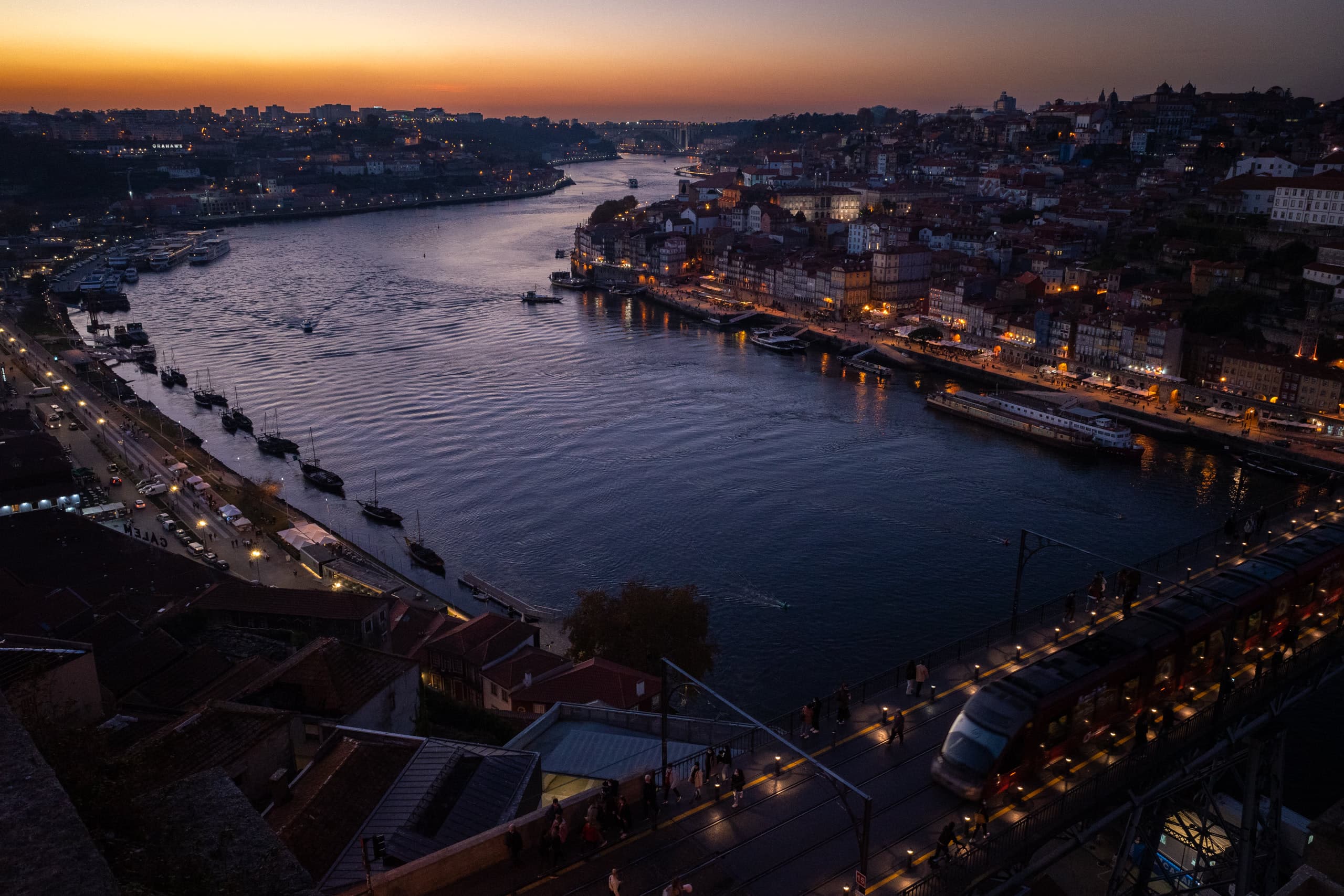 Large river with dense riverfront and a pedestrian bridge at the bottom lit under the sunset