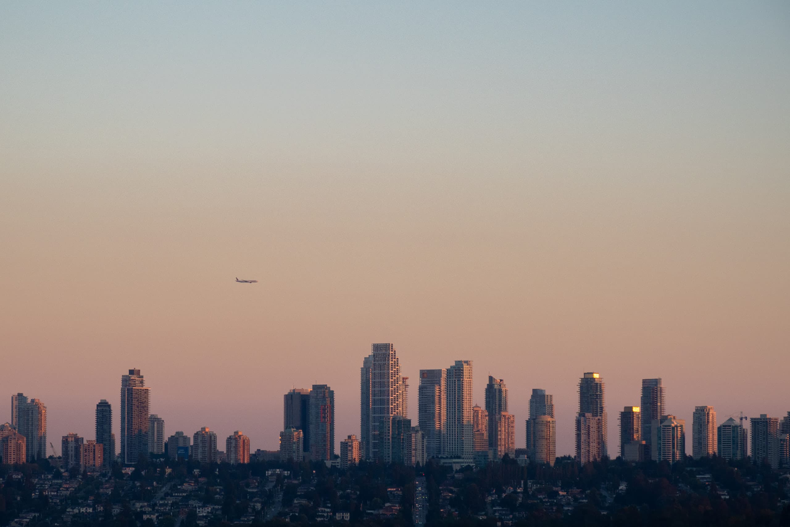 A plane at the left-center flying over a city skyline during sunset