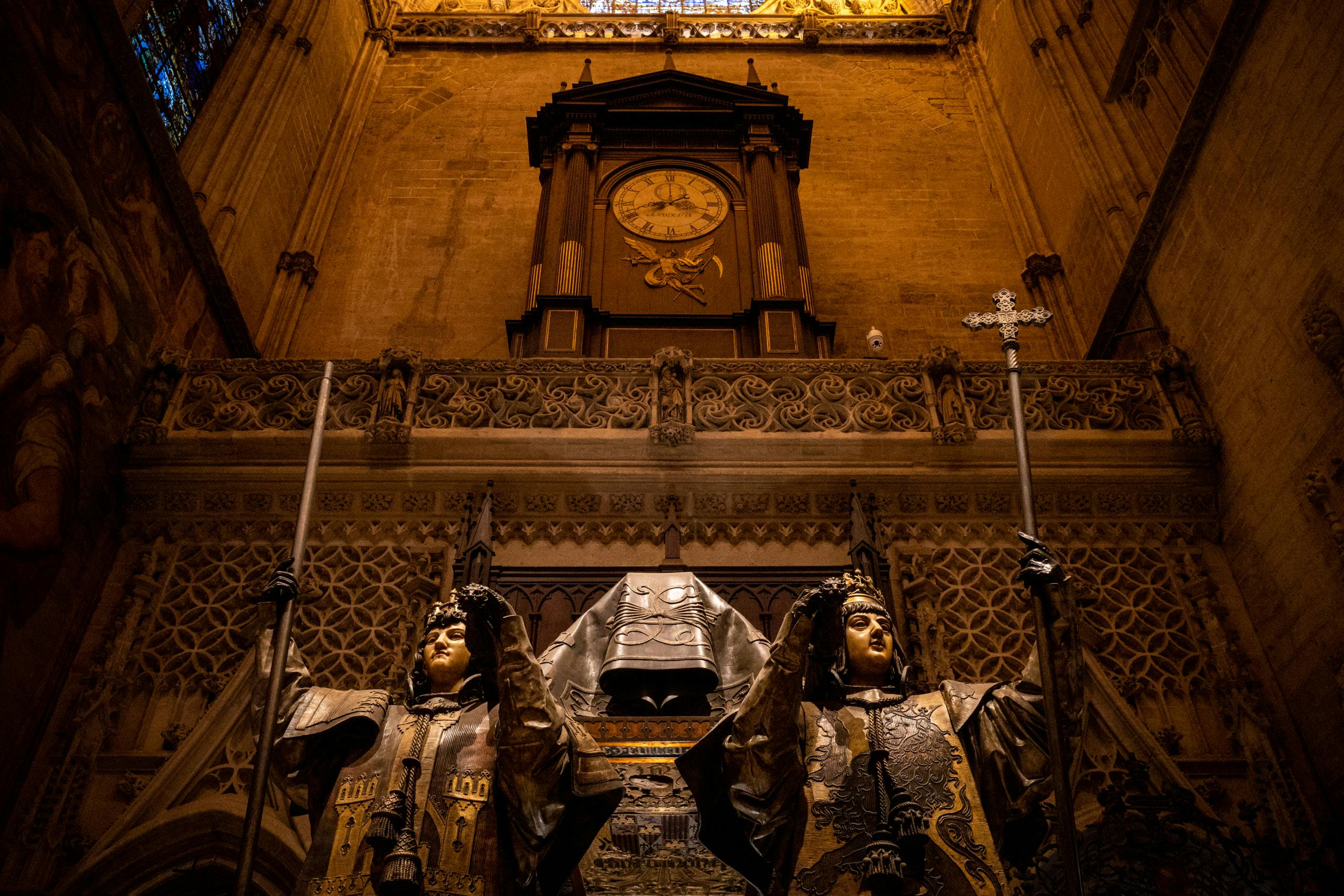 2 statues of priests in front of a holy artifact and a giant clock in the background inside a cathedral