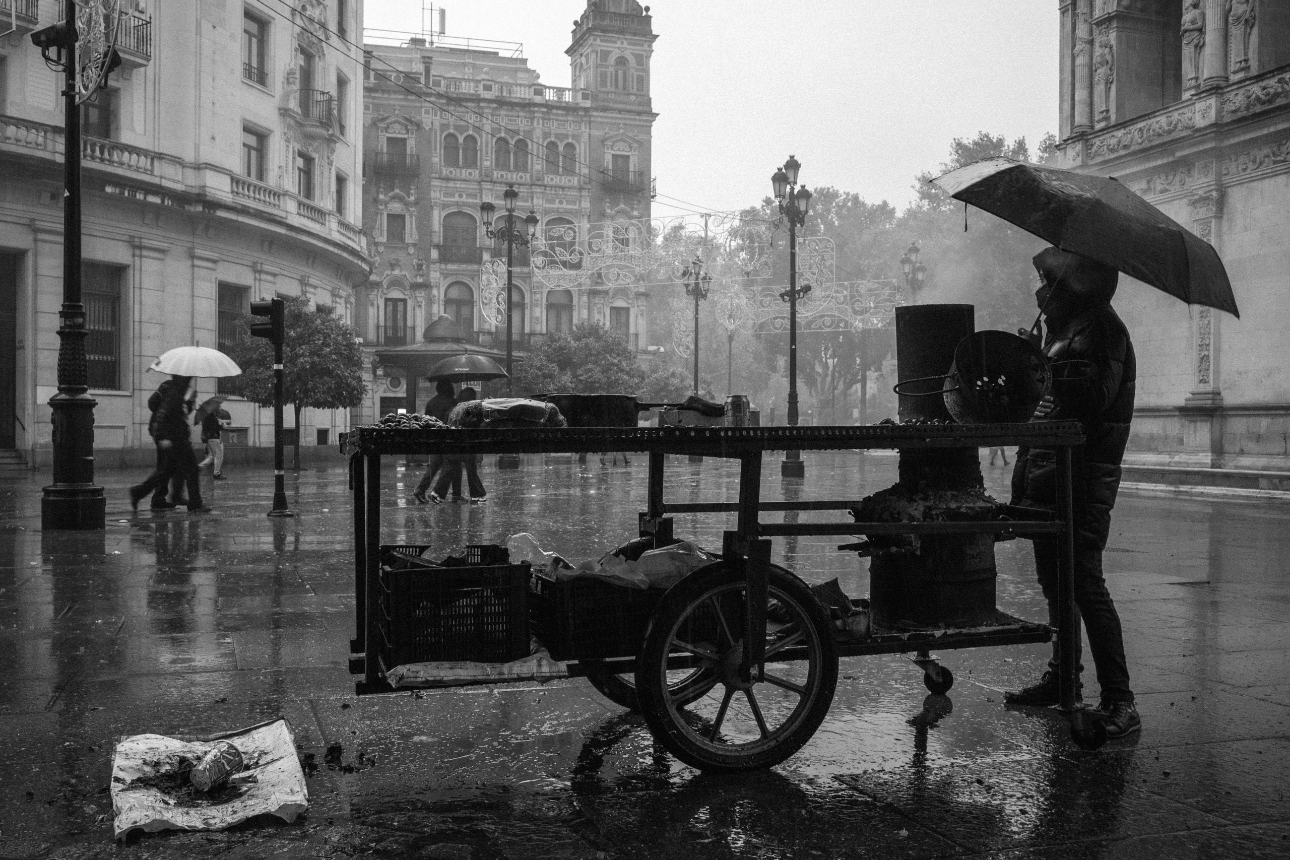 A shop vendor under an umbrella by his cart in the middle of the street