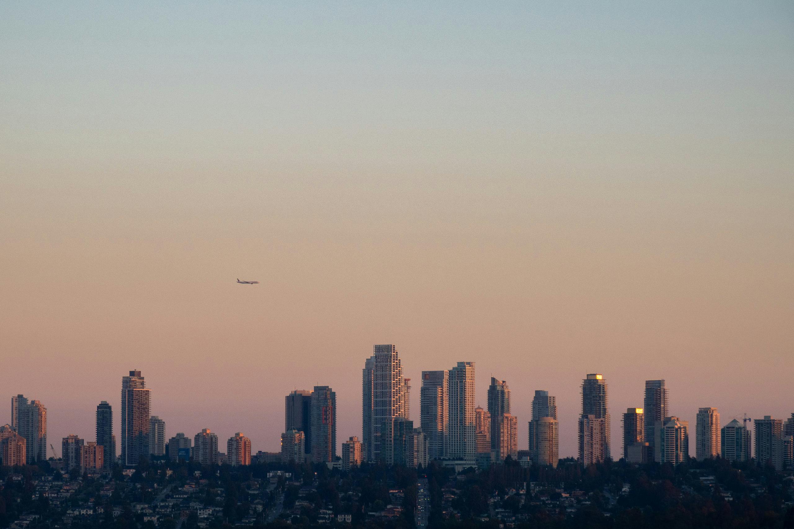 A plane at the left-center flying over a city skyline during sunset